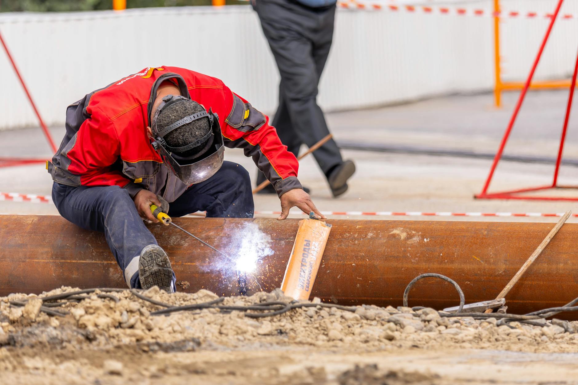 A skilled worker in safety gear welds a metal pipe at a construction site outdoors.
