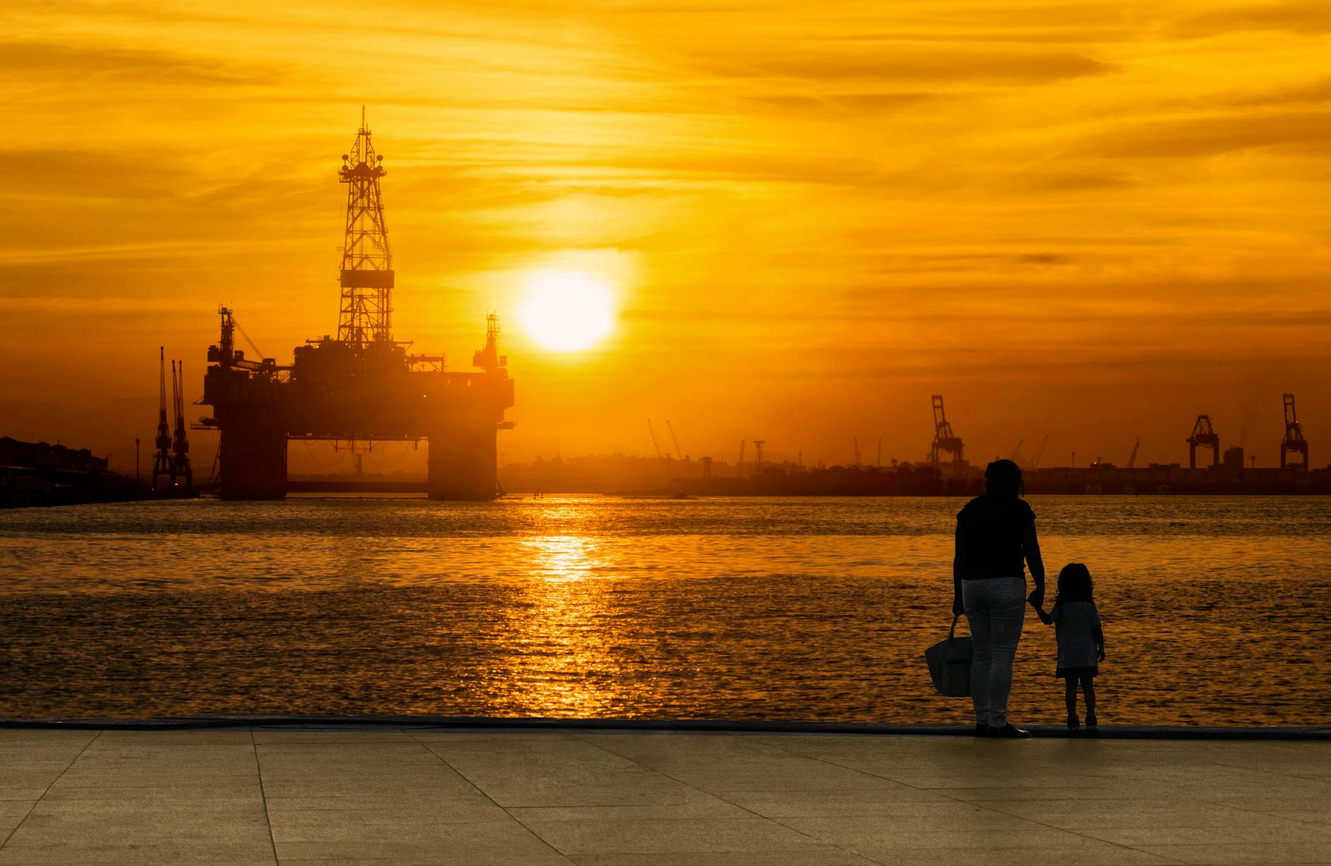 A heartwarming silhouette of a parent and child holding hands at sunset by an oil rig in Rio de Janeiro.