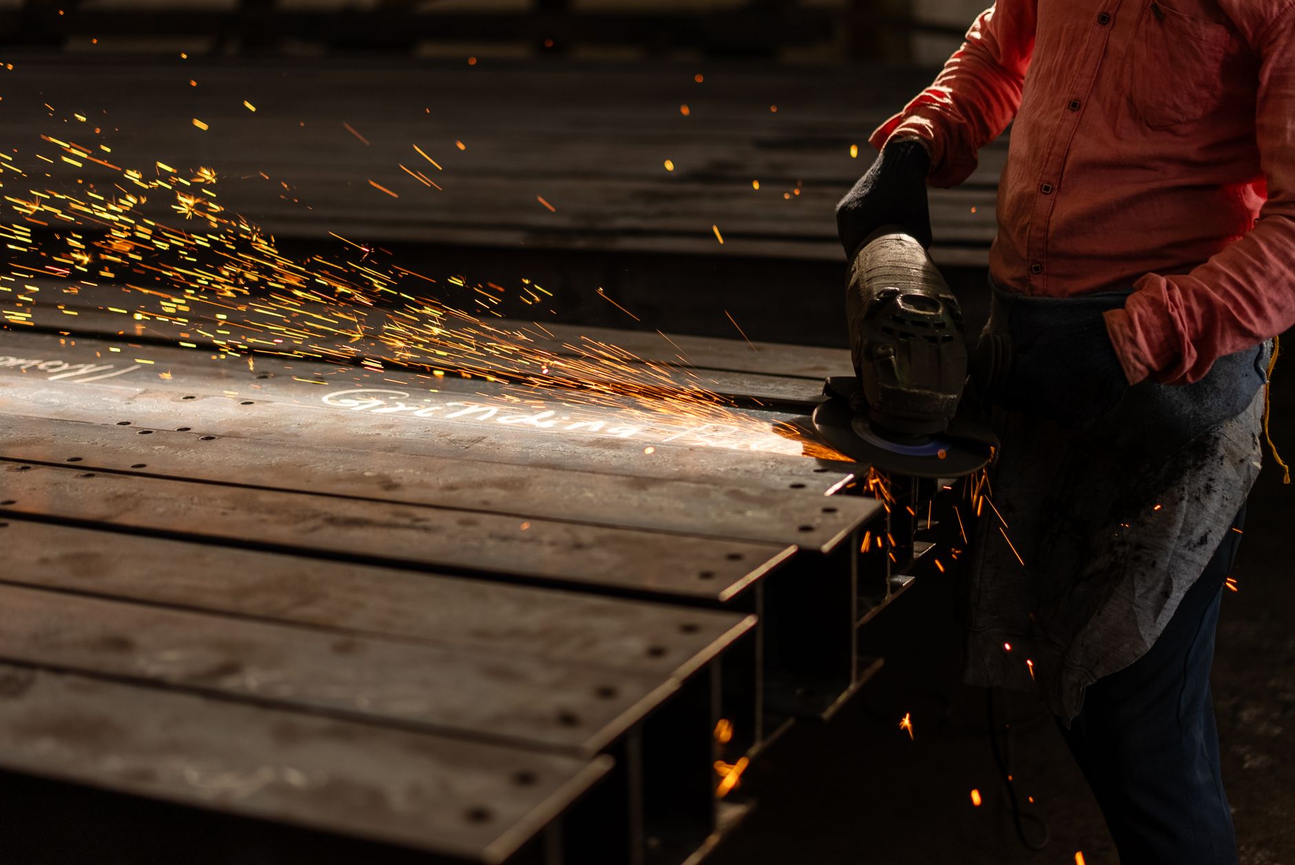 A worker grinds metal beams with an angle grinder, creating vibrant sparks in an industrial setting.